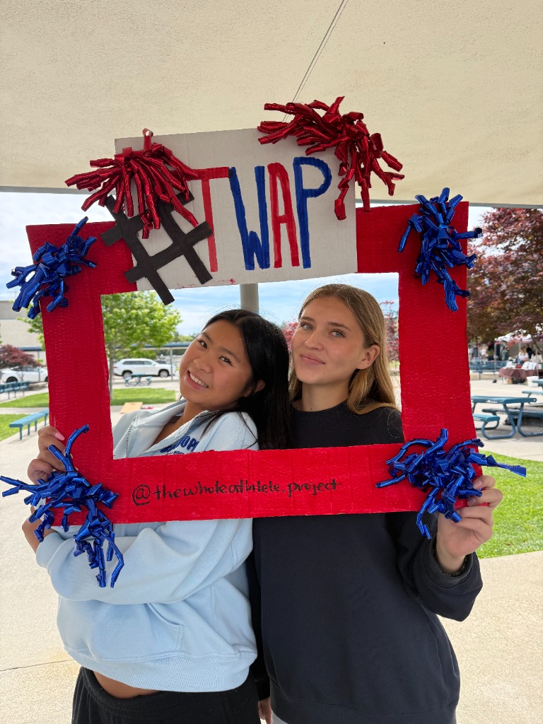 Two student-athletes smiling through a TWAP photo frame at a campus event