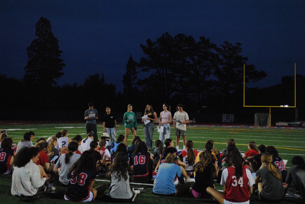 TWAP members presenting to a crowd of student-athletes sitting on a football field at night
